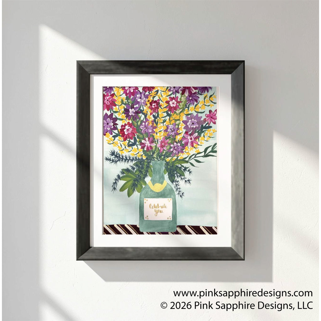 This floral painting features an abundant wildflower bouquet in shades of purple, magenta, and golden yellow, arranged in a sage-green Champagne bottle with a gold foil collar. A small label on the bottle reads “Celebrate You” in gold script, set against a soft mint background above a dark striped tabletop.  The painting has a white mat and black frame and is hung on a white wall.  The text overlay in the bottom right corner say, "www.pinksapphiredesigns.com, ©2026 Pink Sapphire Designs, LLC"