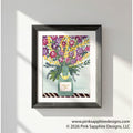 This floral painting features an abundant wildflower bouquet in shades of purple, magenta, and golden yellow, arranged in a sage-green Champagne bottle with a gold foil collar. A small label on the bottle reads “Celebrate You” in gold script, set against a soft mint background above a dark striped tabletop.  The painting has a white mat and black frame and is hung on a white wall.  The text overlay in the bottom right corner say, "www.pinksapphiredesigns.com, ©2026 Pink Sapphire Designs, LLC"