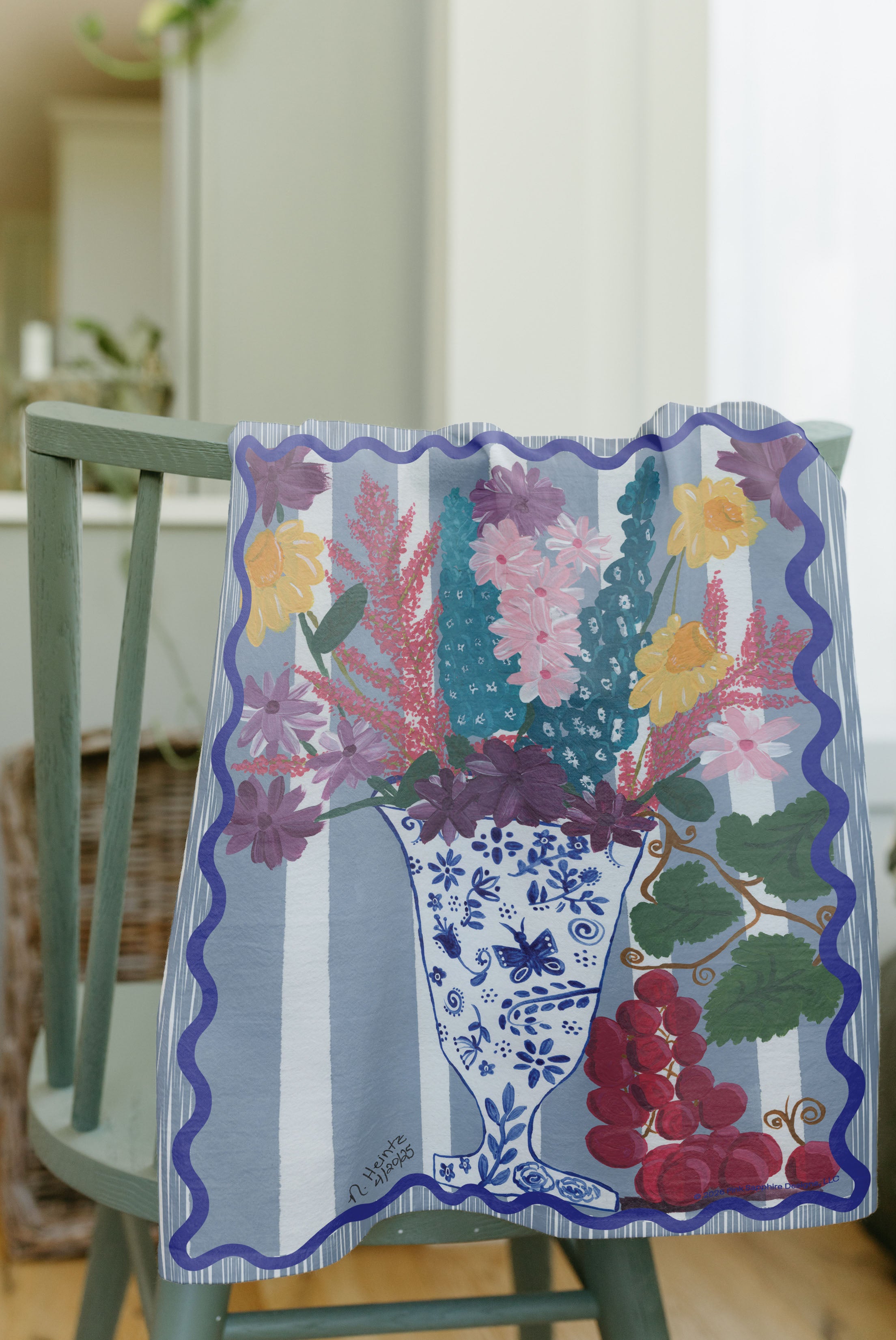 A tea towel draped over a green wooden chair, featuring a colorful floral bouquet in a blue and white patterned vase with  red grapes, set against a light blue and white striped background.