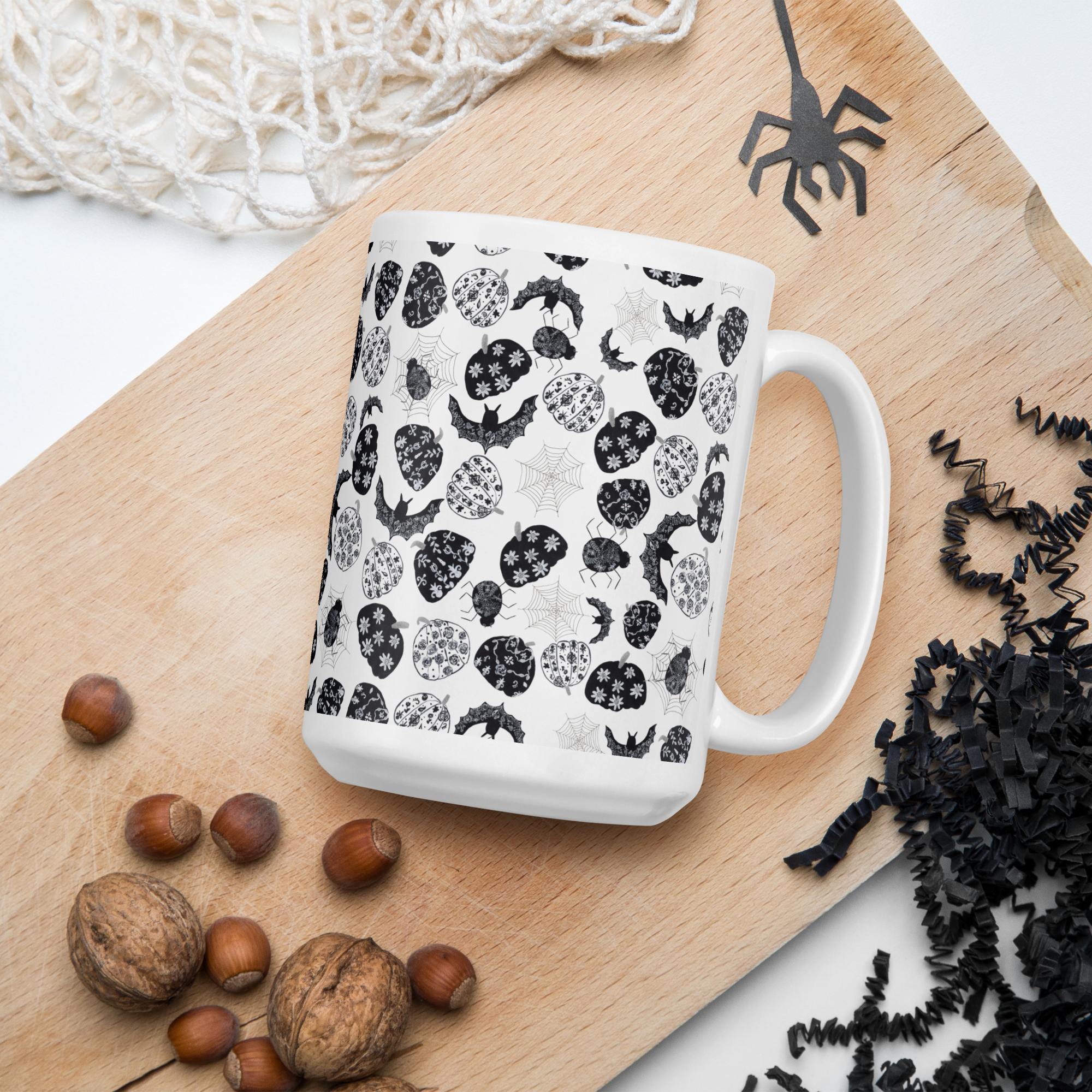 A white ceramic mug with a black and gray Halloween-themed pattern of bats, pumpkins, ghosts, and spiderwebs, styled on a wooden board with nuts and black paper shred.