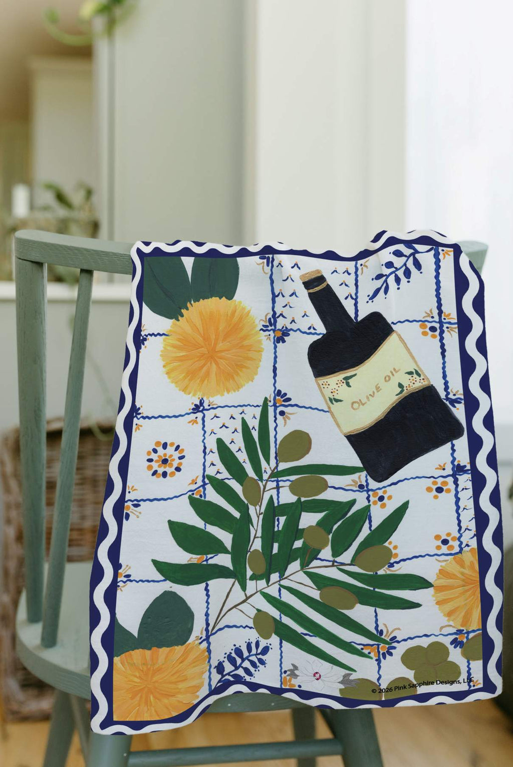 A rectangular textile with a Mediterranean-style print featuring a bottle of olive oil, yellow sunflowers, and olive branches on a blue and white tiled background, draped over a light green wooden chair.