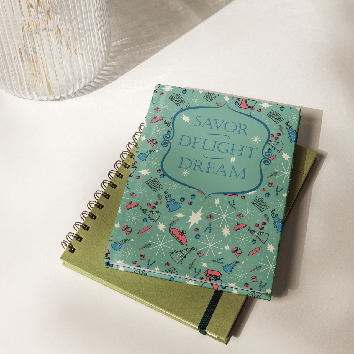 Closed Savor, Delight, Dream hardcover journal stacked on top of a green spiral notebook