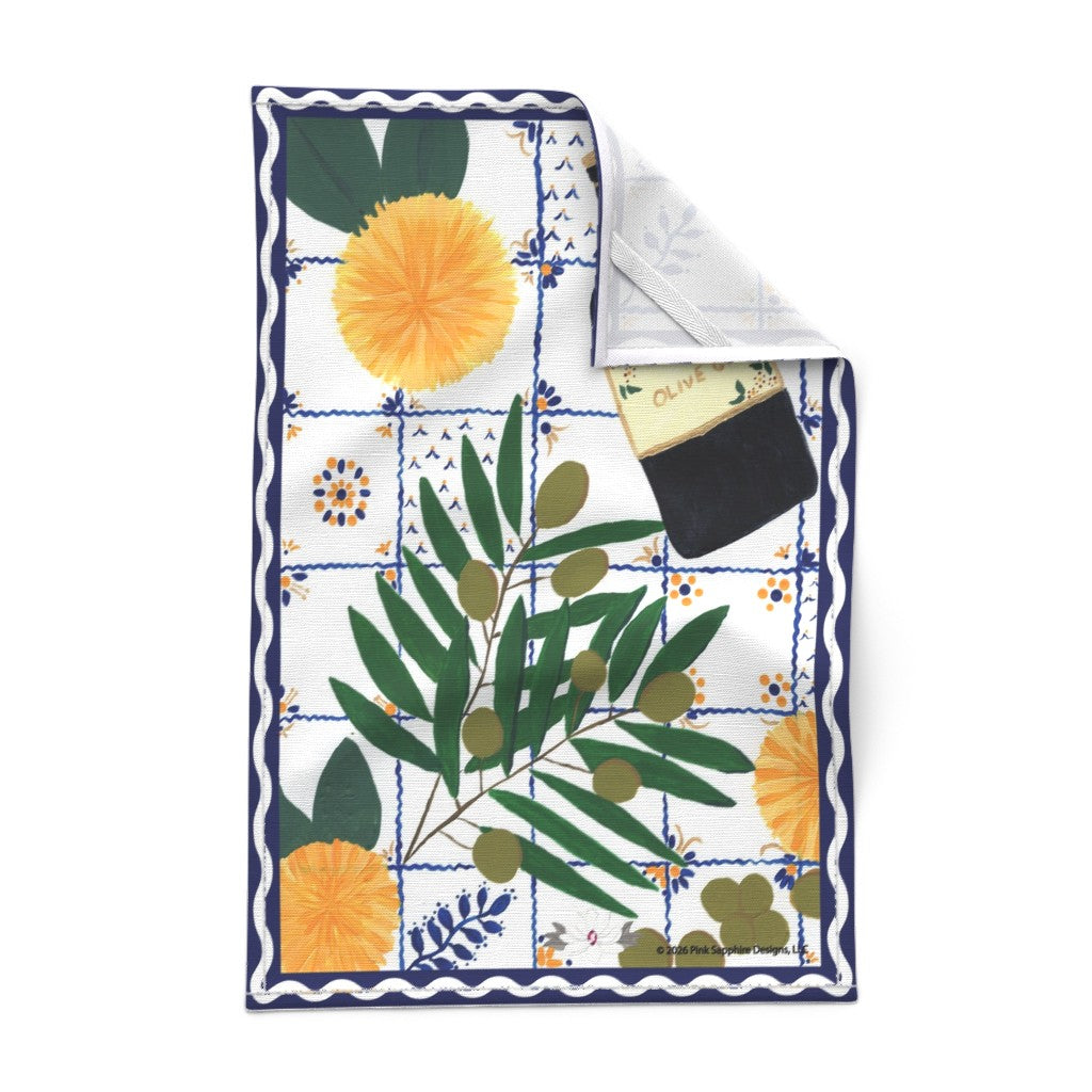 A square kitchen towel with a Mediterranean tile pattern, featuring green olive branches, yellow sun flowers, and blue floral details on a white background. The top-right corner is folded over to show the white textured reverse side and a small tab for hanging the tea towel.